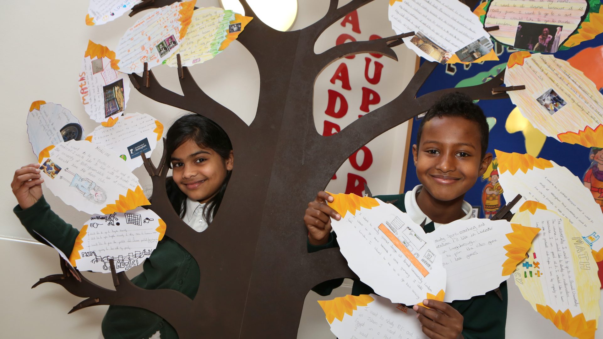 Nottingham Primary students pose near a constructed wooden tree which displays paper leaves where students have written their ambitions for the future.